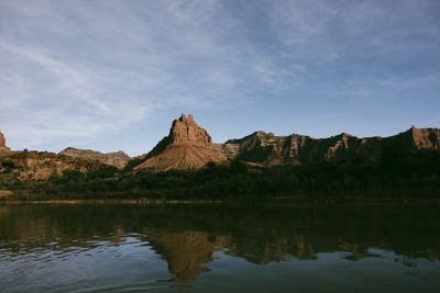 A rock spire and canyon walls reflected in glassy calm water.