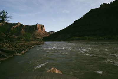 Riffled water runs downstream past a rocky bank toward a sunlit red butte, dark canyon slope opposite.