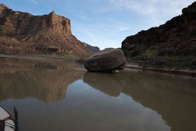 A massive boulder sitting mid-river in glassy water, a red sandstone tower behind.