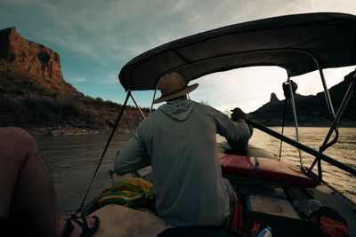 A boater in a straw hat sits under a shade canopy as a second boat runs ahead, red cliffs in evening light.