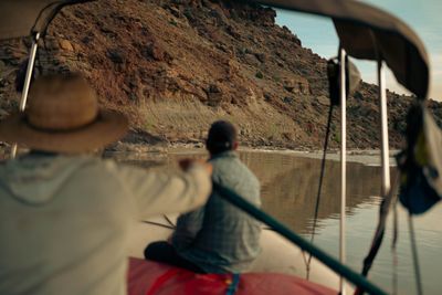 A boater sits on the tube watching a steep layered cliff pass, calm water reflecting the rock.
