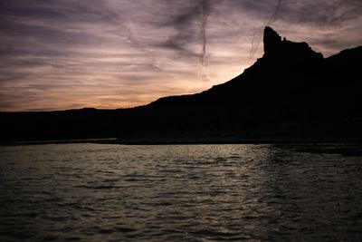 A butte silhouetted against an orange and purple sunset above rippled water.