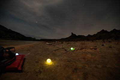 Camper asleep in a sleeping bag on a sandy beach at night, glowing light orbs scattered across the sand, with the river and a dark butte under the stars.