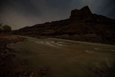 Long-exposure night image of the river flowing silver past a canyon tower under streaking stars.