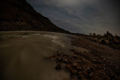 Moonlit long exposure smooths the river past a cobble bank, stars and a bright streak overhead.