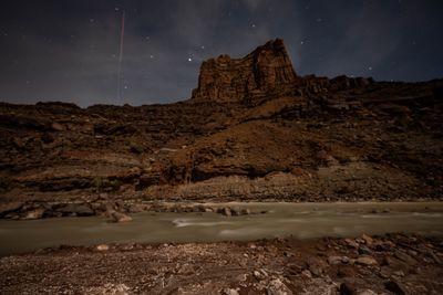 Sandstone tower lit by moonlight with star trails beginning overhead and the river flowing silver below.