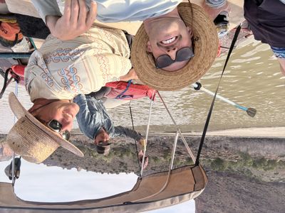 Three crew members in sun hats and sunglasses crowd into a selfie under the raft's shade canopy on flat brown water.