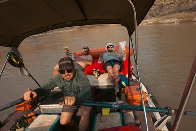 Full crew on the raft, oarsman rowing while three crew members lounge with books and drinks.