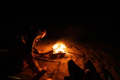 Crew member feeding a branch into the campfire, boots and sand in the foreground.