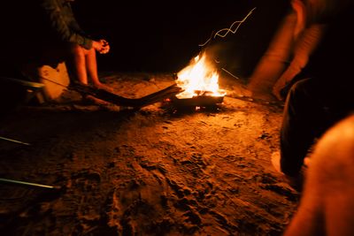 Crew warming hands around a beach campfire, glow sticks on the ground and sparks rising.