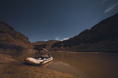 A loaded raft parked on a beach under moonlight, canyon walls faintly lit and stars overhead in a long exposure.