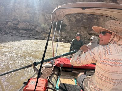 View from the oars of a gear raft: the boatman in a straw hat rows under a shade canopy toward a riffle along a boulder-strewn bank.