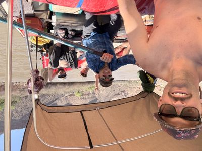 Wide selfie under the raft's shade canopy — four crew grinning around the oar, red dry bags and a badlands bank behind.