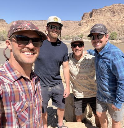 Four crew members pose on a rocky bank above the muddy Green River, cliff-banded mesa walls rising behind.