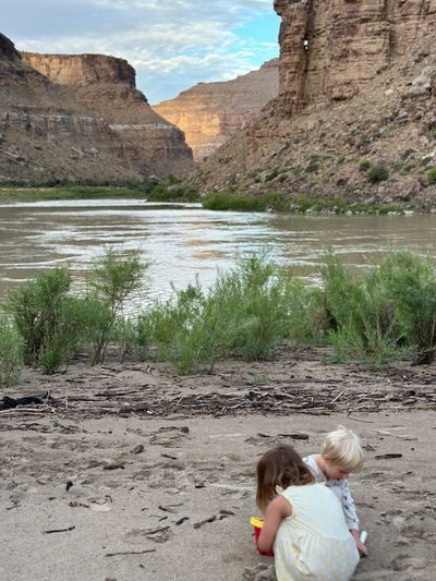 Two small children crouched together digging in the sand of a driftwood-strewn river beach, with green streamside brush, silty brown water, and layered canyon walls rising behind them.
