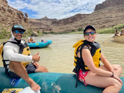 Two crew members in yellow life jackets and sunglasses sitting on the tube of a teal raft, one giving a thumbs-up, with a second raft and a rowed boat drifting behind on silty green water below banded canyon walls.