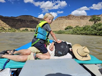 A young child in a blue-and-green life jacket kneeling on a raft's gear pile beside a reclining adult whose straw sun hat rests on the frame, with green streamside brush and stepped canyon walls beyond under a blue sky full of cumulus.