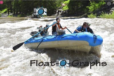 A blue oar raft runs broadside through a silty rapid, four crew aboard in PFDs and ball caps, one hauling an oar high across the frame and a black paddle blade dipped at the left, with green willows and dark bedrock on the bank behind.