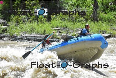 A blue oar raft climbs a breaking wave in a Snake River rapid, the rower leaning back at the stern and a second crew member forward driving a blue paddle into the water, with a rock shelf, deadfall and dense green willows on the bank behind.