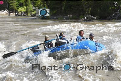 A blue oar raft runs in profile through a long train of silty standing waves on the Snake River, four crew aboard in caps and sunglasses, the upstream oar extended out over the water and a dark conifer bank with driftwood behind.
