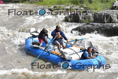 A fully rigged blue oar raft sits in the tail waves of a Snake River rapid seen from above, the rower standing at the oars in an orange PFD, a crew member crouched forward on the tube and two more at the stern, with dark stepped bedrock ledges along the right bank.
