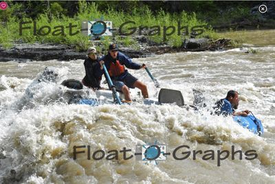 A blue raft is swamped and tipping in a Snake River rapid, two crew braced at the oars with mouths open and the frame seats lifting free, while a third person is in the water at the right holding onto the blue tube, brown foam filling the lower half of the frame.