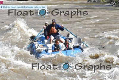 A blue oar raft comes head-on down a wide brown rapid on the Snake River, the rower in a navy jacket and orange PFD pulling both oars, a crew member behind him and two more riding the bow tube as silty standing waves break around the boat.