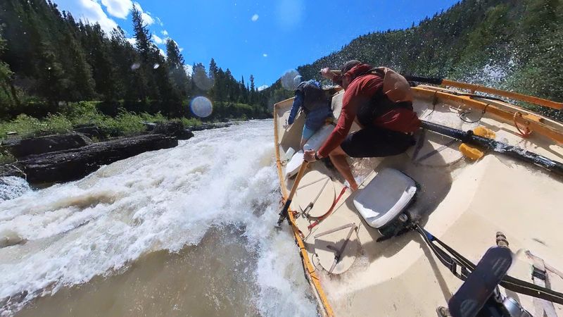A boater in a rust-red shirt and grey cap leans hard into an oar with the blade buried in whitewater beside a wooden dory