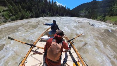 A boater in a blue shirt and cap sits turned around in the bow of a wooden dory pointing downstream, while the rower in a rust-red shirt with a buff pulled over his face holds both oars level, the wide river running fast and riffled between forested slopes.