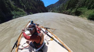A bearded boater in a rust-red long-sleeve shirt, brown wide-brim hat and sunglasses looks back over his shoulder from the oars of a white wooden dory drifting a wide, silty grey-green river, with a second boater in blue seated in the bow and steep conifer-covered canyon walls closing in ahead.