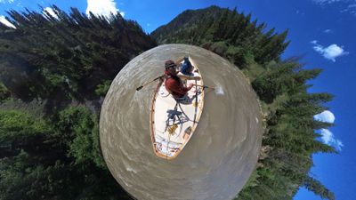 A tiny-planet 360 projection showing a white wooden dory with two boaters aboard floating at the centre of a circular disc of muddy river water, the conifer forest and canyon slopes wrapped radially around the edge beneath deep blue sky and scattered cumulus.