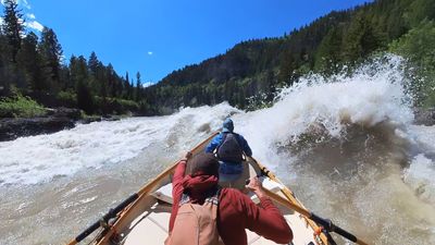 Bow-forward view from a white wooden dory as it drops into a breaking lateral wave that curls across the right side of the frame, the rower in a red shirt and brown hat reaching on the oars and a boater in a blue jacket and teal cap seated low in the bow, forested ridges and blue sky above.