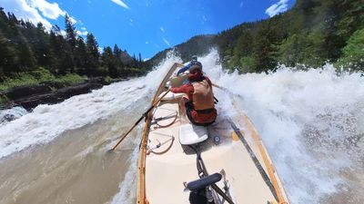 A white wooden dory heeled hard over to the left with a wall of aerated whitewater breaking above the right gunwale, the rower pulling an oar deep in the foam and a boater in blue braced ahead of him, dark bedrock and green brush along the far bank.