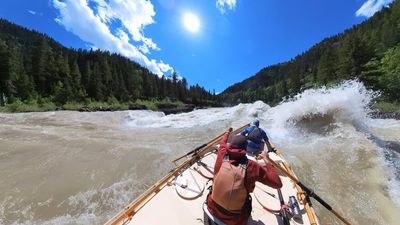 View downriver over the bow of a wooden dory toward a broad line of breaking waves spanning the channel, the rower in red reaching forward on the oars and a boater in a blue jacket seated in the bow, tall conifer ridges on both sides and cumulus building against a deep blue sky.