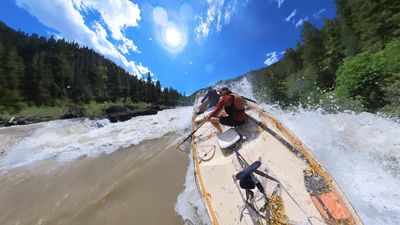 A wooden dory leaning into a field of foaming whitewater with sun flare across the top of the frame, the rower crouched low over the oars and a boater in blue barely visible ahead of him, dark rock ledges along the left bank and forested slopes behind.