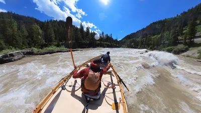 A raised wooden oar blade stands vertically in the foreground of a wooden dory running a long wave train, the rower seated below it in a rust-red shirt and a boater in blue in the bow, forested canyon walls on both sides and a road grade cut into the slope on the right.