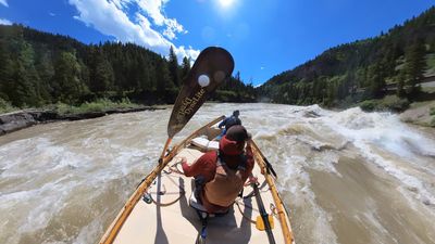 A wooden oar blade swings across the upper frame of a bow-forward view from a dory in a wave train, the rower hunched over the oars in a rust-red shirt and a boater in blue seated ahead, conifer slopes on both banks and a road grade on the right hillside.