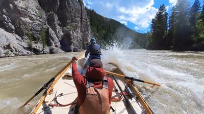 A wooden dory runs beside a tall grey cliff face on the left bank while a burst of whitewater breaks over the right side of the boat, a boater in a blue jacket standing braced in the bow and the rower's arm raised on the oar, conifer slopes and cumulus cloud beyond.