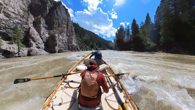 Bow-forward view from a wooden dory on smooth silty water beneath a tall grey rock face on the left bank, a boater in blue leaning over the bow and the rower seated at the oars, forested ridges and towering cumulus opening downstream.