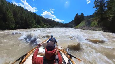 A wooden dory sits centred in a broad train of breaking waves on a wide brown river, the rower's arms spread on the oars and a boater in a blue jacket and teal cap in the bow, forested slopes on both sides and scattered cumulus across a deep blue sky.
