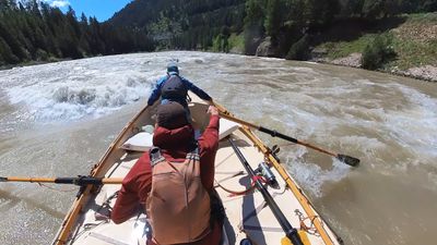A boater in a rust-red shirt and tan life vest reaches forward on an oar aboard a wooden dory while a second boater in blue leans out over the bow, breaking waves stacked along the left side of the channel and a gravel and brush bank on the right.