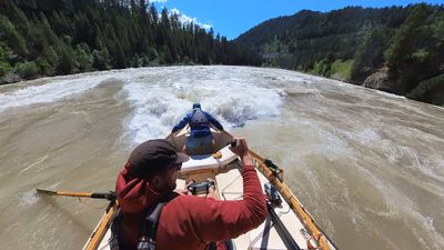 A bearded boater in a brown cap and sunglasses works the oars of a wooden dory, face turned partly toward the camera, while a second boater in a blue jacket crouches over the bow as the boat enters a broad line of breaking waves on a fast brown river.