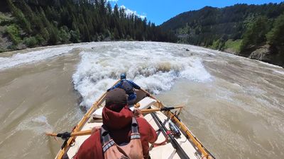 A wooden dory noses into a large curling wave that fills the middle of the channel, whitewater rising level with the gunwale, a boater in a blue jacket and teal cap braced in the bow and the rower pulling hard on the oars, forested canyon slopes beyond.