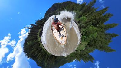 A tiny-planet 360 projection showing a wooden dory with two boaters punching a large breaking wave that arcs across the top of a circular disc of river, conifer forest wrapped around the rim beneath blue sky and cumulus.