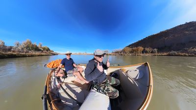 Two men float a wooden drift boat down a calm, muddy-green river with autumn cottonwoods lining the far bank and a bare sandstone hillside on river right under a clear blue sky.