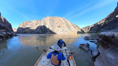 Two boaters in a wooden dory drift across a calm green pool in Westwater Canyon, a sunlit sandstone-capped rock face rising ahead above dark inner-gorge walls under a clear blue sky.