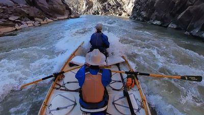 Wooden dory runs a whitewater rapid down the center of a tight dark-walled gorge in Westwater Canyon, both boaters leaning into breaking waves.
