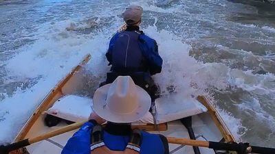 Close view from the stern of a dory as breaking whitewater washes over the deck, two boaters hunched forward in the froth.