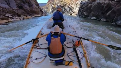 Wooden dory drops into a whitewater rapid deep in Westwater Canyon, spray flying as both boaters push through, dark schist walls squeezing the river.