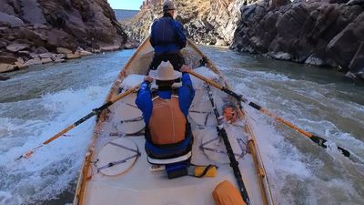 Boaters power a wooden dory into a large breaking wave in a Westwater Canyon rapid, whitewater surging over the deck between sheer dark walls with a boulder on river right.